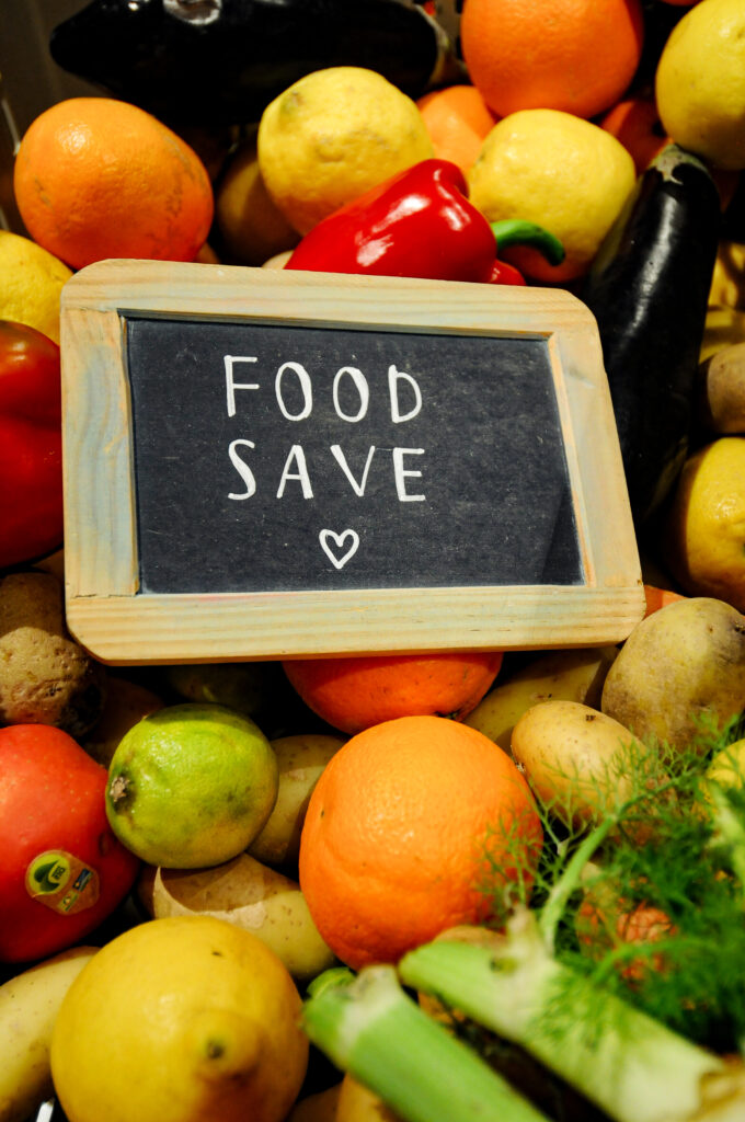 Ein Schild mit der Aufschrift "Food Save" liegt in einem Korb gefüllt mit Früchten und Gemüse. 